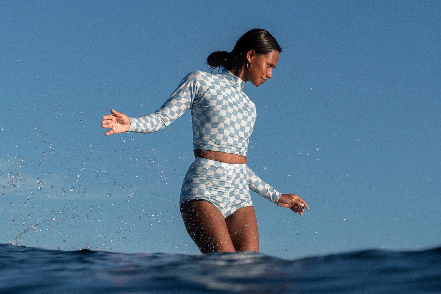 Woman surfing on a longboard wearing a white and blue checkered two-piece set from SunDaze Surf