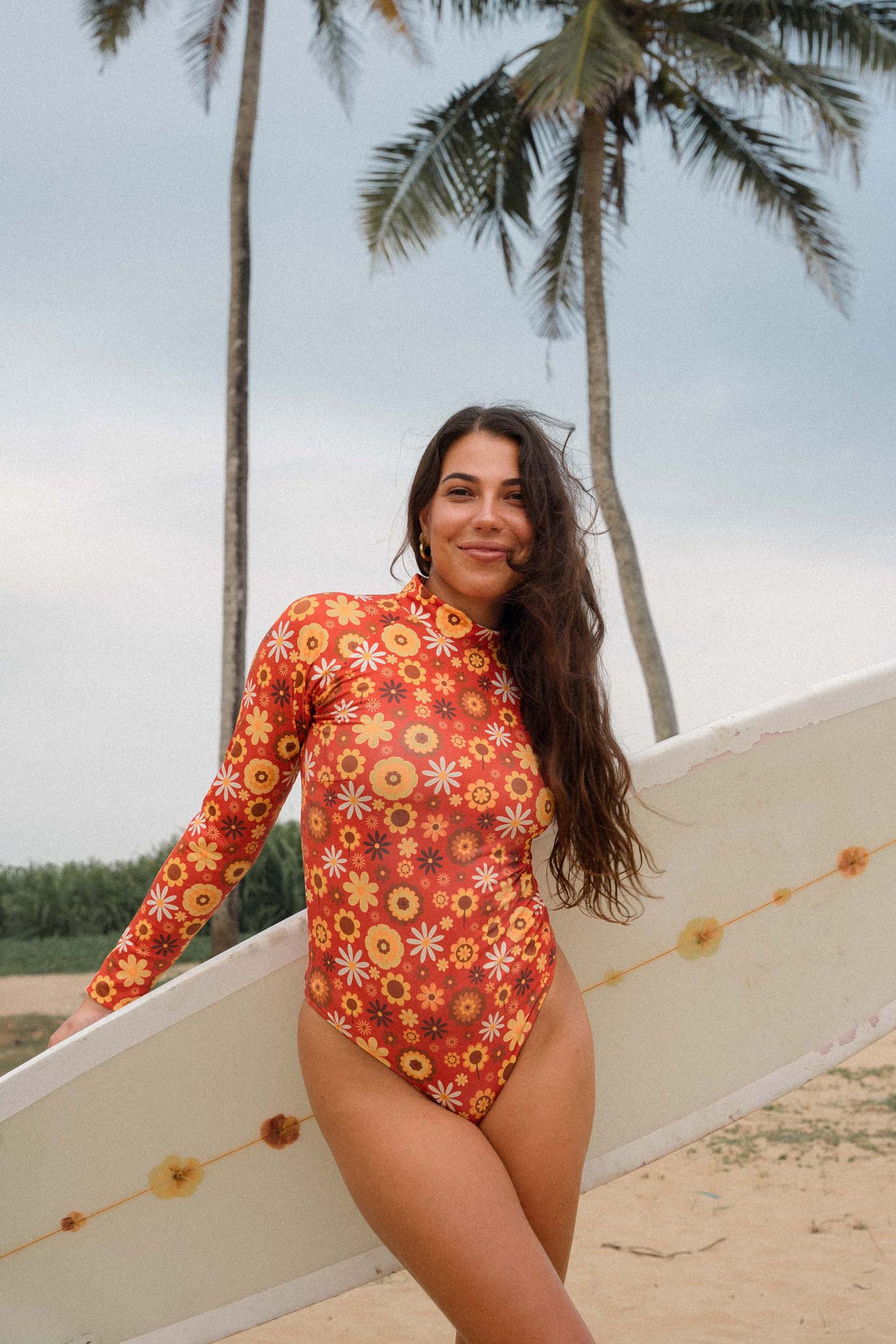 Model posing at the beach with a surfboard wearing the long sleeve surf suit with retro floral print