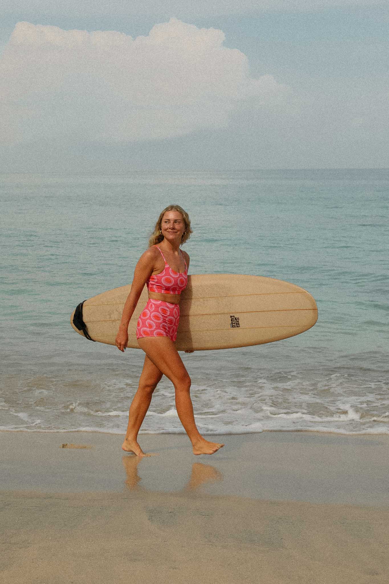 model walking on the beach with her surfboard wearing the watermelon bikini