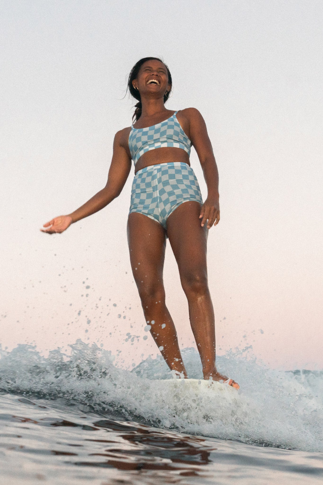 Woman surfing and smiling broadly with the sunset in the back, wearing the putu blue check set