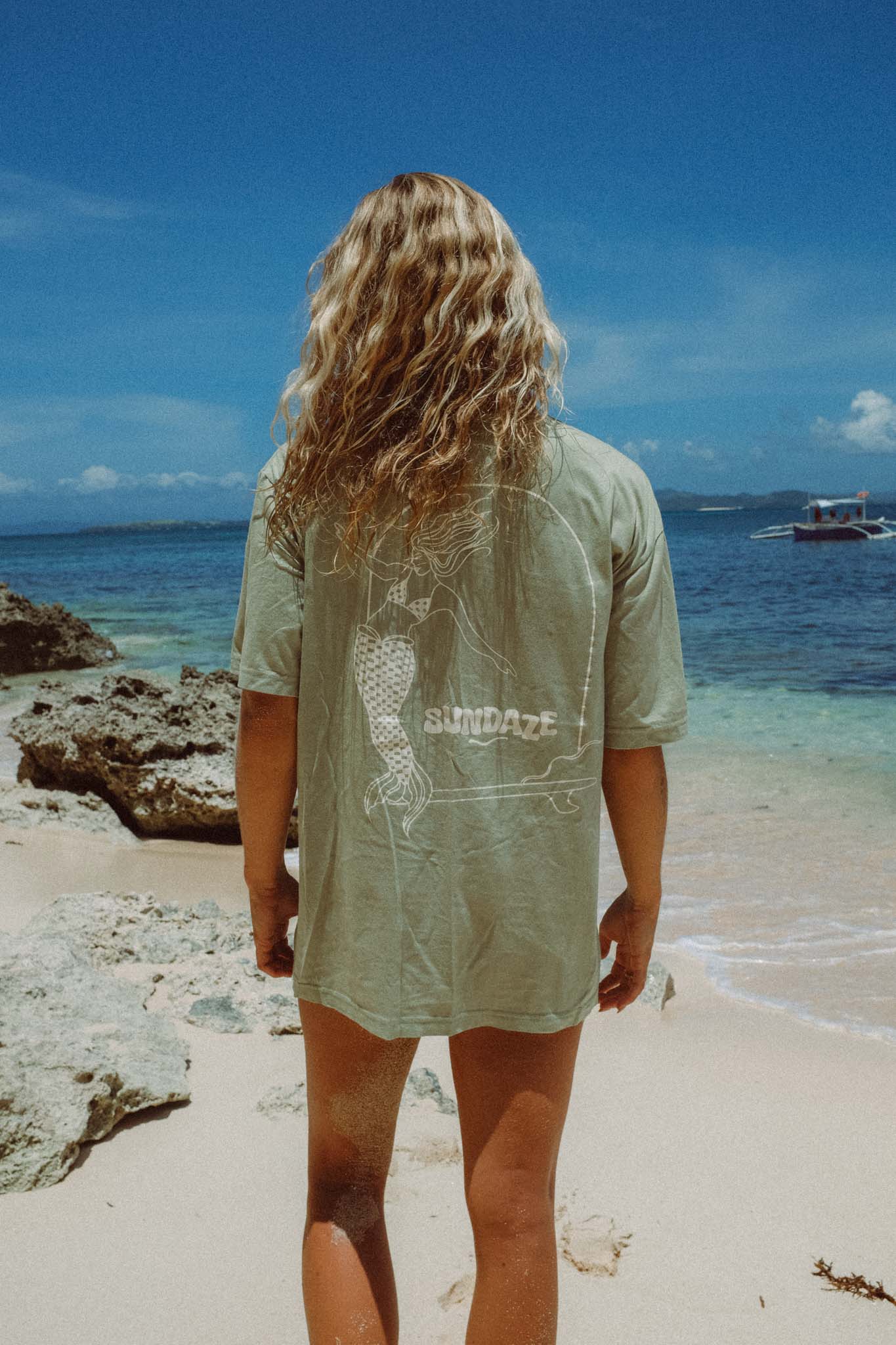 Model at the beach wearing a sun hat and showing the back of her t-shirt, a sage green t-shirt with mermaid illustration